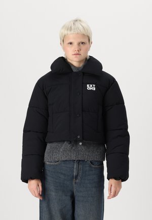 Black cropped puffer jacket with large collar, button closure, and white "EXT" logo. Worn over a gray sweater, features oversized puffed sleeves.