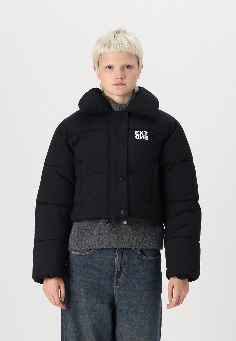 Sort cropped pufferjakke med stor krave, knaplukning og hvid "EXT" logo. Bæret over en grå sweater, med overdimensionerede, puffede ærmer.