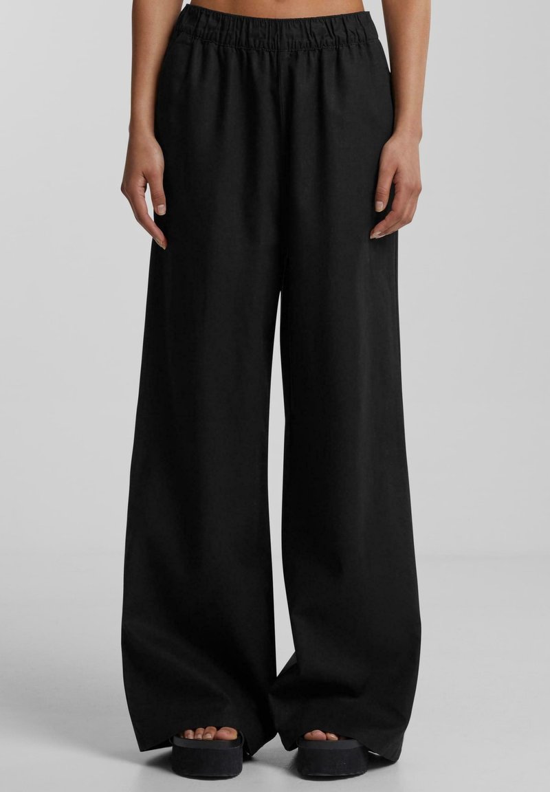 Pantalon noir à jambes larges avec taille élastique, porté avec des sandales à plateforme noires sur pieds nus, visible de la taille jusqu'au sol.