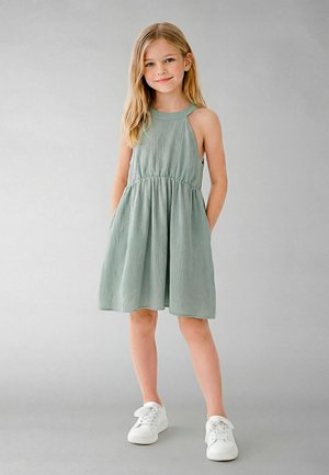 Ung jente med langt blond hår iført en ermeløs salviegrønn kjole og hvite sneakers, stående foran en ensfarget grå bakgrunn.