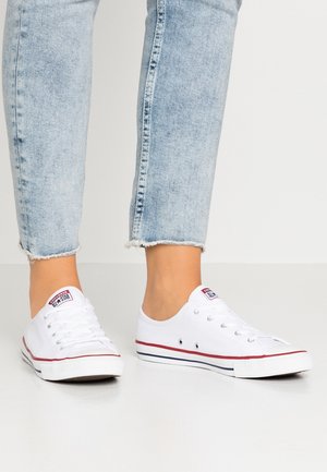 Personne portant un jean court bleu clair et des baskets blanches basses Converse All Star debout sur un sol blanc.