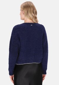 Kobieta z blond włosami ubrana w ciemnoniebieski puszysty sweter i czarną spódnicę, pokazana od tyłu na jasnym tle.