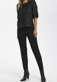 Kvinna som bär en svart texturerad puffärmstop, åtsittande svarta byxor och svarta klackade ankelstövlar, stående mot en vit bakgrund.