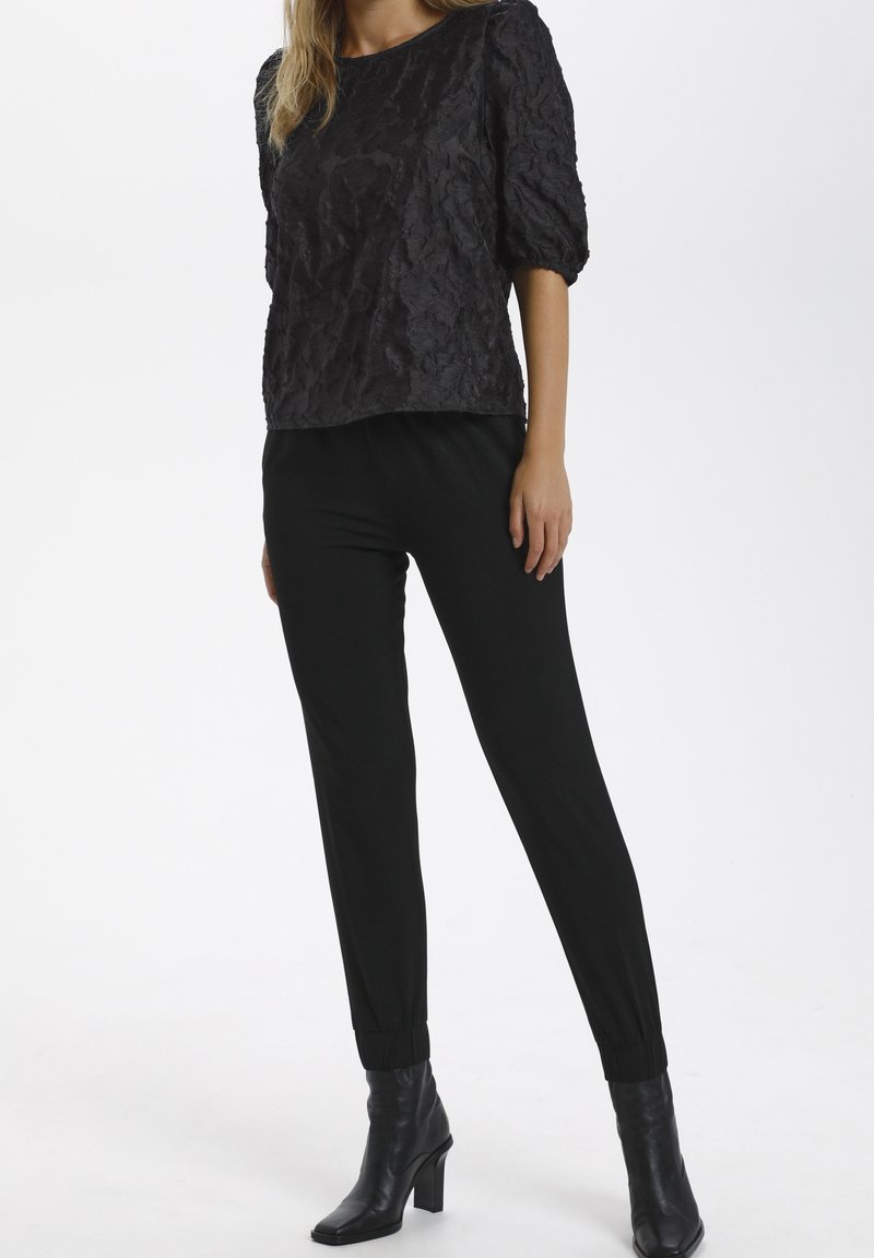 Kvinna som bär en svart texturerad puffärmstop, åtsittande svarta byxor och svarta klackade ankelstövlar, stående mot en vit bakgrund.