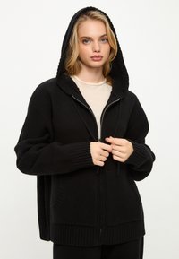 Černá zipová mikina vyrobená z měkkého materiálu. Má žebrovaný lem, velké kapsy a kapuci se šňůrkou. Hladká textura a volný střih.