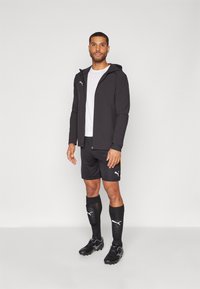 Black ribbed jacket with a hood, white t-shirt underneath, black shorts with yellow accent, black socks, and soccer cleats.