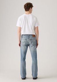 Man wearing a plain white t-shirt and light blue straight-leg jeans, standing with back facing the camera against a neutral background.