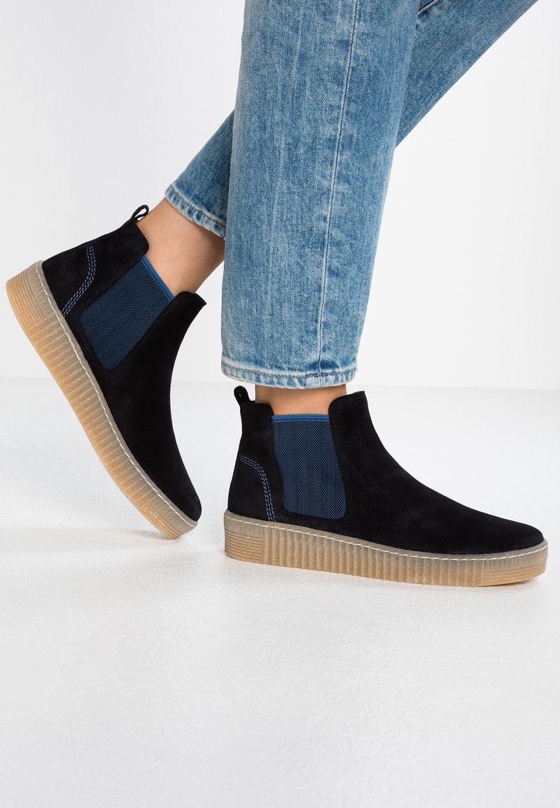 Personne portant un jean en denim bleu et des bottines Chelsea en daim noir avec des côtés élastiques bleus et des semelles en caoutchouc marron clair.