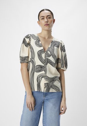 Blusa crema con patrones de líneas abstractas en negro, escote en V pronunciado y mangas abullonadas. Combinada con jeans de color azul claro.