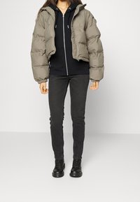 Nainen, jolla on päällään oliivinvihreä lyhyt puffer-takki mustan vetoketjuhupparin päällä, mustat skinny-farkut ja mustat nauhakiinnitteiset saappaat, seisoo yksinkertaisella taustalla.