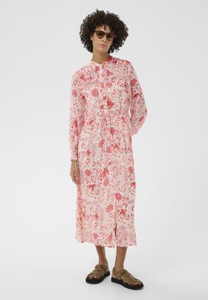 Femme portant une robe rose à manches longues boutonnée avec un imprimé floral rouge et des oiseaux, de grandes lunettes de soleil et des sandales à lanières marron, debout sur un fond blanc.