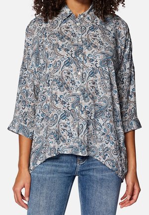 Vrouw draagt een losse blouse met paisley-patroon, 3/4 mouwtjes en een blauwe spijkerbroek.