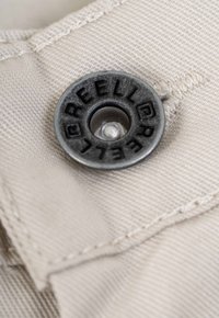 Metal button with engraved "REELL" on a light beige fabric with visible stitching, showcasing a textured surface and smooth finish.
