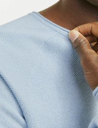 Pulover tricotat de culoare albastru deschis, evidențiind textura și detalii ale cusăturii, cu o mână care ajustează gâtul. Dispune de un guler nervurat și cusături.