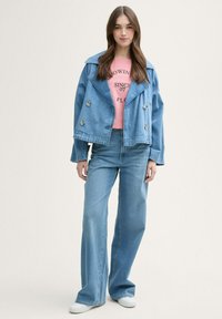 Chaqueta de mezclilla con amplios solapas y botones de doble pecho, combinado con una camiseta gráfica rosa y unos vaqueros de pierna ancha de color azul claro. Zapatos blancos.
