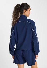 Mörkblå sportjacka med hög krage, långa ärmar och elastiska muddar, i kombination med matchande shorts. Har vit passpoal som detalj.