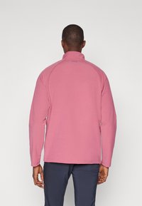 Fleece pulover v mehki roza barvi z visokim ovratnikom, dolgimi rokavi in ravnimi šivi. Ima manjši logotip blizu vratu na hrbtu.