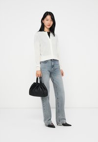 Jeune femme portant un cardigan blanc, un top noir, un jean bleu clair, des chaussures noires à bout pointu, et tenant un sac à main noir froncé.