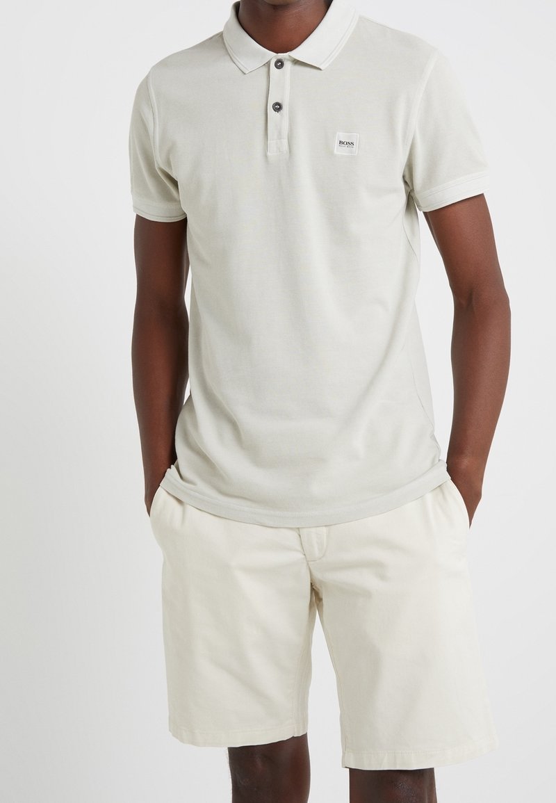 Homme portant un polo beige clair à manches courtes avec deux boutons et un short beige assorti, les mains dans les poches.
