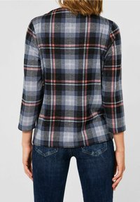 Nainen, joka on pukeutunut pitkähihainen ruutupaita mustan, sinisen ja vaaleanpunaisen sävyissä yhdistettynä tummansinisiin farkkuihin, nähtynä takaa.