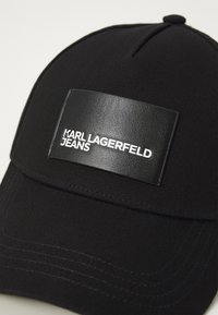 Crna kapa od teksturirane tkanine s pravokutnim kožnim uzorkom na kojem je utisnut bijeli natpis "KARL LAGERFELD JEANS."