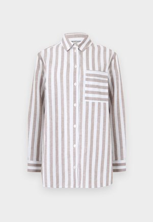 Nő bézs és fehér, függőleges csíkos, laza ingben és hozzá illő, bő szárú nadrágban, fehér felsővel alatta, egy kézzel a zsebében áll.