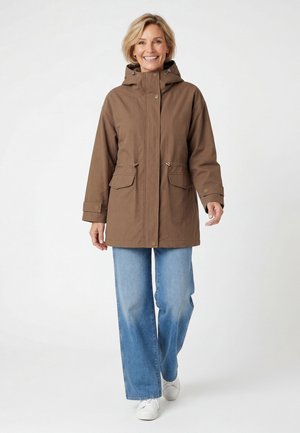 Smiling woman walking wearing a brown hooded jacket, light blue wide-leg jeans, and white sneakers on a plain white background.