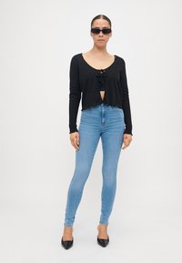 Svart kortermet cardigan med knyting foran, kombinert med lyseblå smale jeans og svarte spisse hæler. Tilbehør inkluderer mørke solbriller.