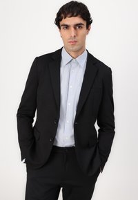 Schwarze, maßgeschneiderte Sakkjacke über einem hellblauen Hemd mit Knopfleiste. Die Jacke hat zwei Knöpfe und ein elegantes Design. Sauberer, minimalistischer Look.