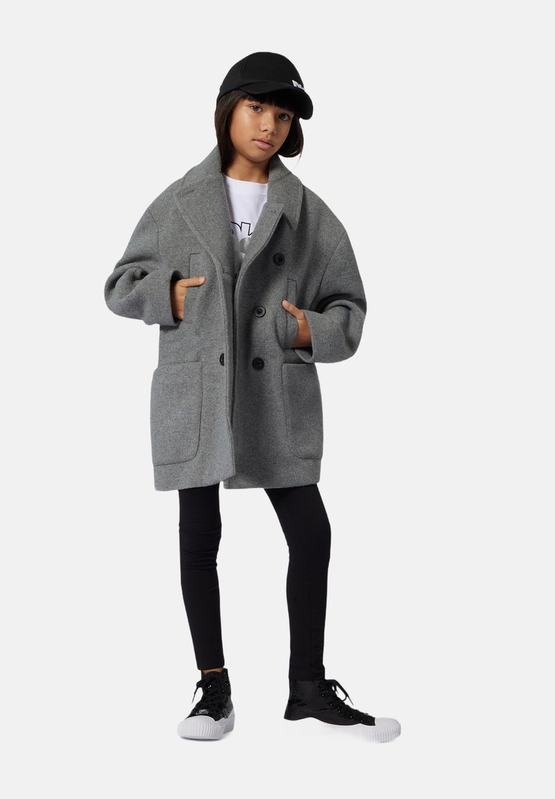 Niño con un abrigo gris de doble botonadura, leggings negros, gorra negra y zapatillas altas negras, de pie con las manos en los bolsillos.