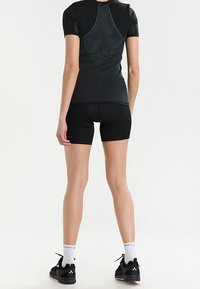 Person wearing black short-sleeve athletic shirt and padded cycling shorts, standing with back facing the camera against a white background.