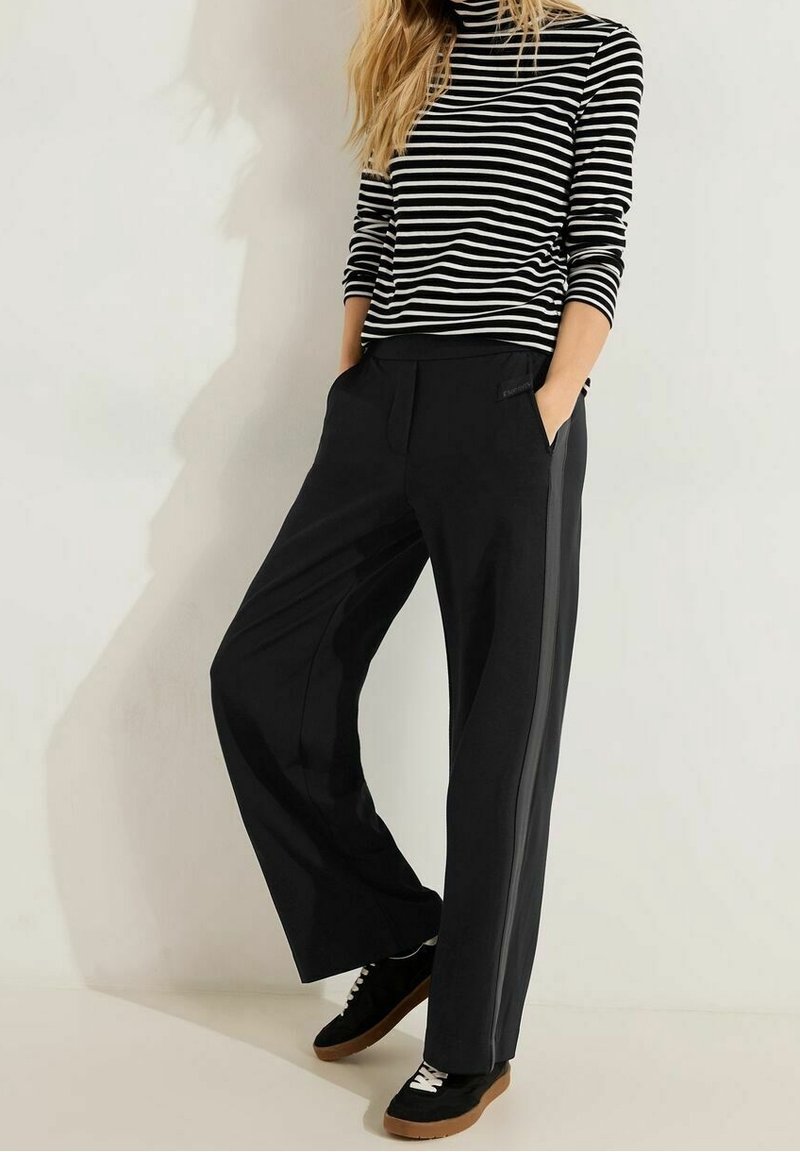 Femme portant un pantalon large noir, un haut à manches longues rayé noir et blanc, et des baskets noires à lacets blancs et semelles en gomme, debout les mains dans les poches.
