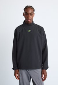 Black pullover jacket with high collar, elastic cuffs, and green logo. Smooth fabric, slightly oversized fit, minimalistic design.