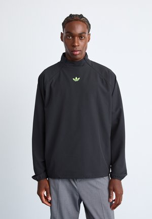Black pullover jacket with high collar, elastic cuffs, and green logo. Smooth fabric, slightly oversized fit, minimalistic design.