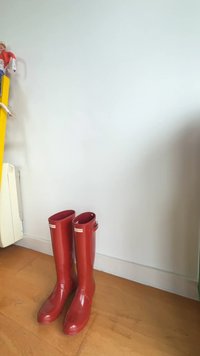 Red rubber rain boots with a glossy finish, featuring a rounded toe and adjustable strap accents. Positioned on a wooden floor against a wall.