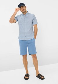 Lichtblauw kortarmhemd met een geometrisch patroon, gecombineerd met lichtblauwe shorts en zwarte sandalen, met een donkere pet.