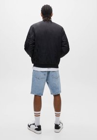 Blouson en cuir noir avec des poignets côtelés, porté avec un short en jean bleu clair. Des chaussettes blanches à rayures marron et des baskets noires complètent le look.