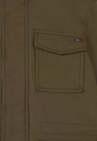 Olivově zelená bunda s přední kapsou s klopou, vyšitými detaily a malým značkovým emblémem v červené, bílé a modré barvě.