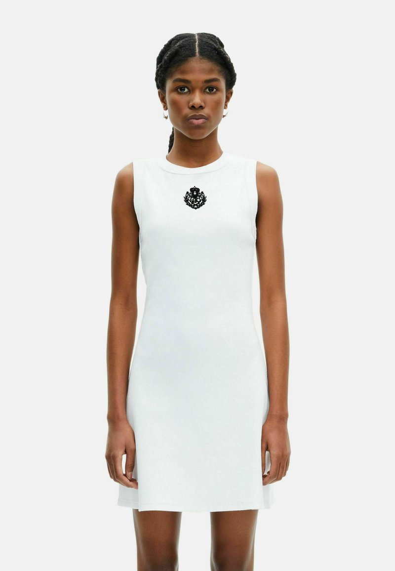Donna con i capelli intrecciati che indossa un vestito bianco senza maniche con un emblema nero centrato sul petto, in piedi contro uno sfondo semplice.