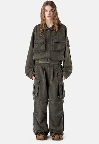 Persona joven con cabello largo que lleva una chaqueta cargo verde oscuro de gran tamaño y pantalones anchos a juego con múltiples bolsillos, de pie mirando hacia adelante.