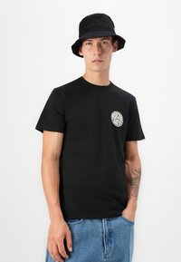 Svart bomulls t-skjorte med rund hals og korte ermer, med en hvit sirkulær logo på venstre bryst. Kombinert med lyse denimshorts.