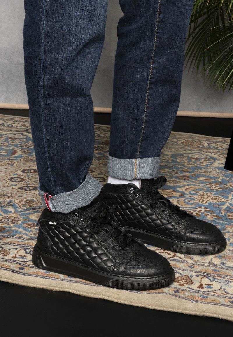 Black high-top sneakers with quilted texture, lace-up design, and rubber sole, paired with rolled-up dark denim jeans and white socks.