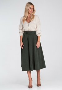Jasno beżowy sweter z dzianiny z guzikami; ciemnozielona spódnica z wysokim stanem z paskiem i kieszeniami; gładka tekstura; noszona z brązowymi szpilkami.