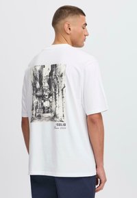 Hvid kortærmet t-shirt med et sort-hvidt grafisk print på ryggen, der viser en sidegade, med teksten "ISOLID Siden 1999."
