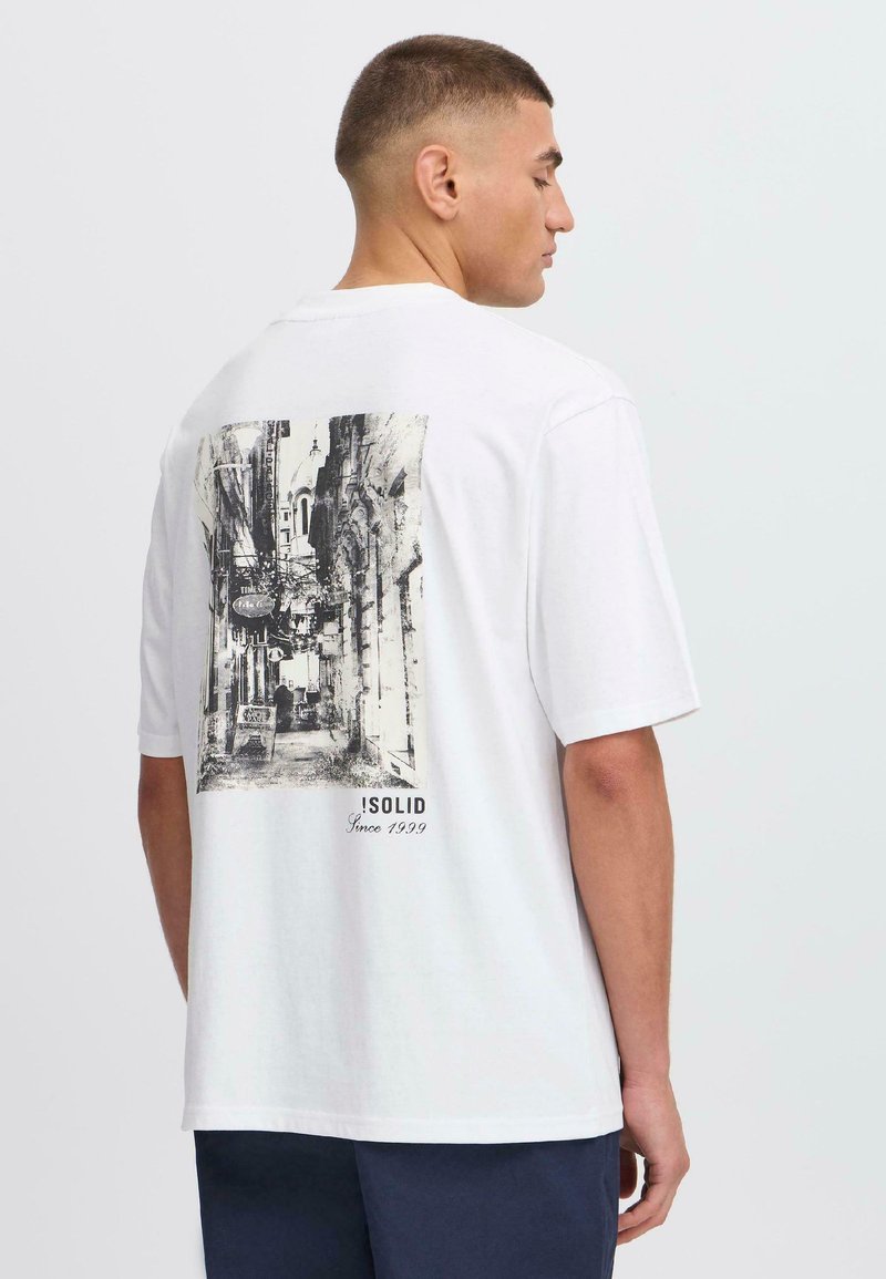 Hvid kortærmet t-shirt med et sort-hvidt grafisk print på ryggen, der viser en sidegade, med teksten "ISOLID Siden 1999."