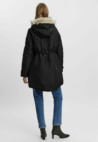 Parka noire isolée avec une capuche doublée de fourrure, taille cintrée et manches longues. Associée à un jean bleu et des bottines à talons noirs.