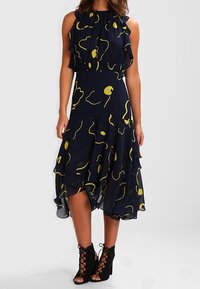 Laivastonsininen mekko röyhelötyllä halterneck-olkaimella, keltainen abstrakti printti ja keinuva A-linjainen siluetti. Pukeutunut mustiin nyörikorkokenkiin.