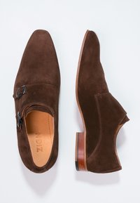 Scarpe in suede marrone con punta affilata e ritagli laterali. Presentano un dettaglio con fibbia e un tacco in legno con una leggera sfumatura.