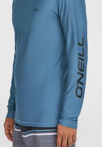 Jasnoniebieski rash guard z długim rękawem z czarnym logo "O'NEILL" na prawym rękawie, wykonany z gładkiego, elastycznego materiału.