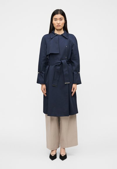 Trench-coat bleu marine avec un col raccourci, un devant croisé, une ceinture à nouer et des manches longues ; associé à un pantalon beige à jambes larges et des talons noirs.